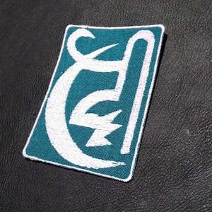Puede incluir: Parche bordado en color turquesa y blanco con un diseño estilizado de la letra "E" y un rayo. El parche es rectangular con un borde blanco y está sobre un fondo negro texturizado.