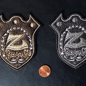 Pode incluir: Dois patches bordados em forma de escudo com uma letra Z estilizada dentro de uma coroa. O patch da esquerda é marrom com bordado dourado. O patch da direita é preto com bordado prateado.