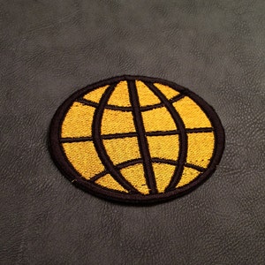 May include: A yellow embroidered globe patch with a black outline. The globe is on a dark grey background.