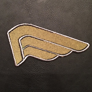 Peut inclure: Un écusson brodé doré en forme d'aile avec un contour blanc.