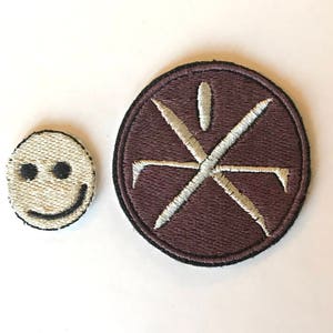May include: Two embroidered patches. One is a small, off-white smiley face with black eyes and a curved mouth. The other is a larger, circular patch with a dark brown background and a white, stylized cross design.