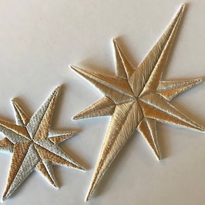 Könnte beinhalten: Zwei gestickte Sternformen, eine in Gold und eine in Weiß, mit einem dezenten Schimmer. Der goldene Stern ist größer und hat eine spitzere Form, während der weiße Stern kleiner ist und eine abgerundete Form hat.