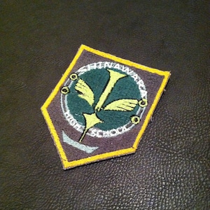 Könnte beinhalten: Ein Stoffaufnäher mit geometrischer Form, der ein zentrales Emblem mit einem vogelartigen Design in Gelb und Grün zeigt. Der Aufnäher trägt den Text "SHINAWARA HIGH SCHOOL" in Weiß, umrandet von einem gelben Rand.