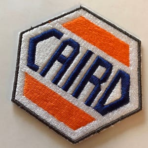 Könnte beinhalten: Ein weißer, sechseckiger Patch mit einem orange-weißen Streifenmuster. Das Wort "CAIRO" ist in Blau auf den Patch gestickt.