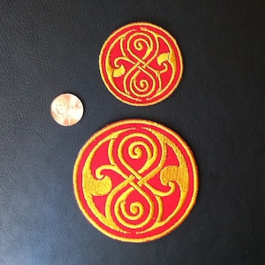 May include: Two embroidered patches with a red background and a gold-colored symbol. The symbol is a stylized infinity symbol with swirls and loops. The patches are circular and are different sizes.