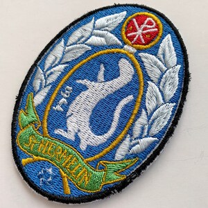 May include: A blue and white embroidered patch with a white stylized animal, a green and gold ribbon with the text "Shroomin", and a red and white symbol in the upper right corner.