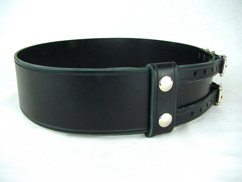 View of left side of black double buckle kilt belt. View features double snap leather keeper and detail design grooves carved in each buckle strap and along edges of belt strap and leather keeper.