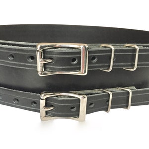 Detail view of front of black double buckle kilt belt. View features nickel plate buckles and two metal keepers per buckle strap. Also seen is the detail design groove carved in each buckle strap.