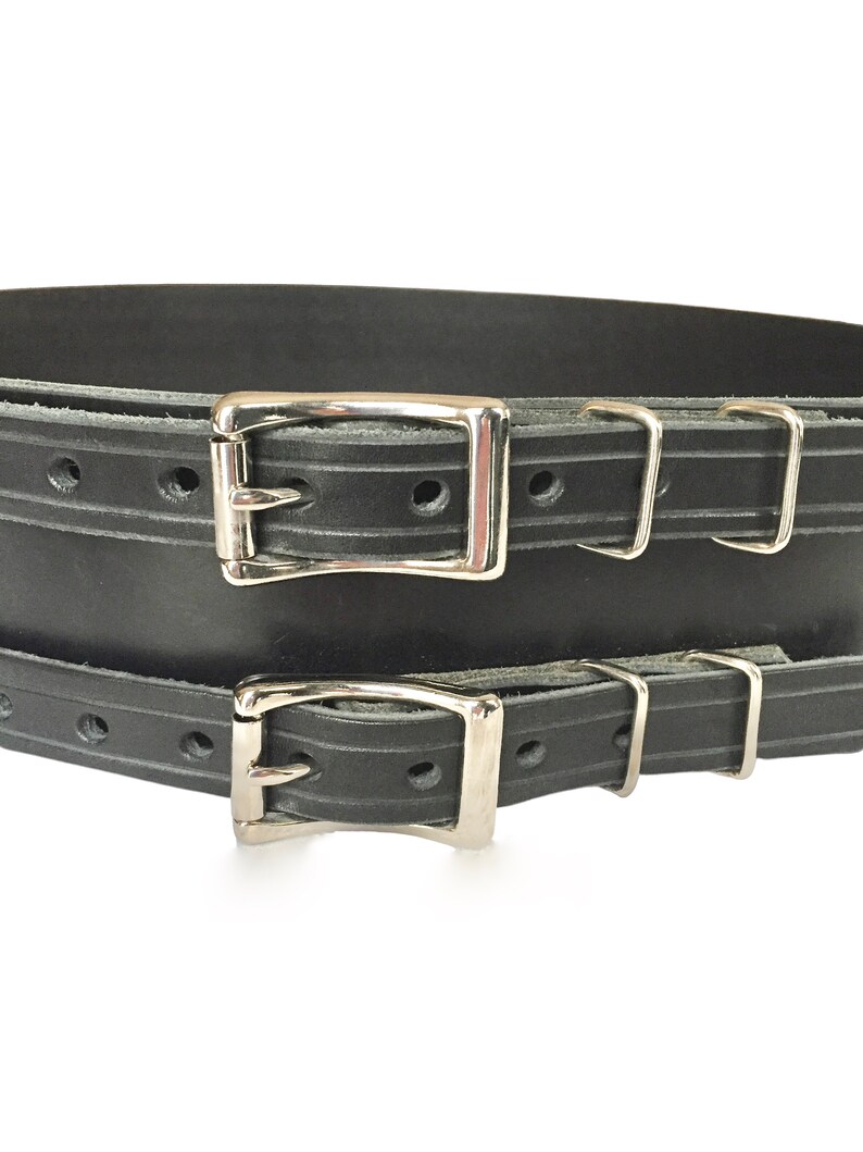 Detail view of front of black double buckle kilt belt. View features nickel plate buckles and two metal keepers per buckle strap. Also seen is the detail design groove carved in each buckle strap.