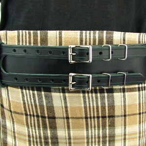 View of front of belt worn with tartan. Black double buckle kilt belt with nickel plate buckles and two metal keepers per buckle strap. Also shown on left is double snap keeper. This keeper holds the overlapping belt leather behind the belt.