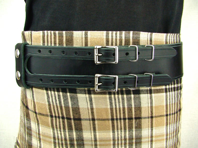 View of front of belt worn with tartan. Black double buckle kilt belt with nickel plate buckles and two metal keepers per buckle strap. Also shown on left is double snap keeper. This keeper holds the overlapping belt leather behind the belt.