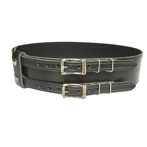 View of front of belt. Black double buckle kilt belt with nickel plate buckles and two metal keepers per buckle strap. Also shown on left is double snap keeper. This keeper holds the overlapping belt leather behind the belt.