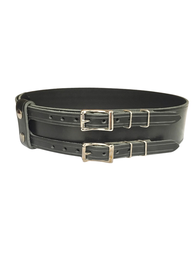 View of front of belt. Black double buckle kilt belt with nickel plate buckles and two metal keepers per buckle strap. Also shown on left is double snap keeper. This keeper holds the overlapping belt leather behind the belt.
