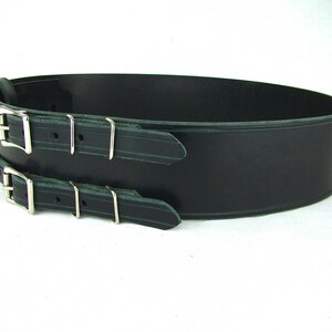 View of right side of black double buckle kilt belt. View features nickel plate buckles and two metal keepers per buckle strap. Also seen is the detail design groove carved in each buckle strap and along edge of belt strap.