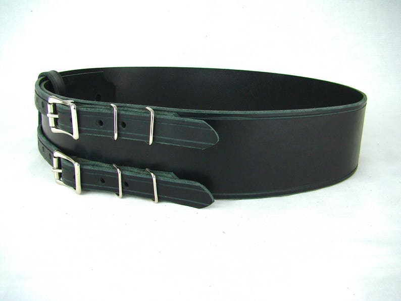 View of right side of black double buckle kilt belt. View features nickel plate buckles and two metal keepers per buckle strap. Also seen is the detail design groove carved in each buckle strap and along edge of belt strap.
