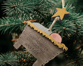 Peut inclure: Ornement de Noël en bois en forme de crèche, avec une étoile dorée et le texte "THE GIFT THAT FOREVER CHANGED THE world." L'ornement a un style rustique et est suspendu à un sapin de Noël.