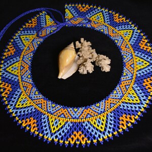 May include: A beaded necklace with a geometric pattern in blue, yellow, and orange colors. The necklace is laid out on a black surface with a seashell and coral.