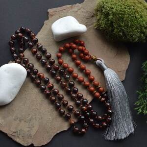 May include: A mala or prayer beads made of red and black stones with a grey tassel. The mala is on a stone surface with a white stone and green moss.