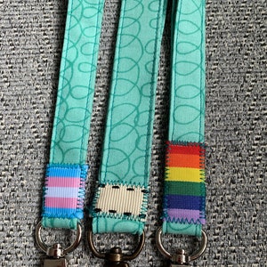 May include: Three teal blue patterned lanyards with different colored fabric patches. The patches are a transgender pride flag, a rainbow pride flag, and a white fabric patch with black stitching. Each lanyard has a silver or bronze metal clip.
