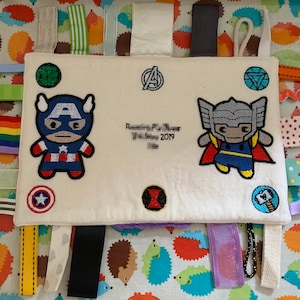 May include: A white fabric baby sensory toy with various textures and colours. It features embroidered images of Captain America and Thor, along with the text "Remembering the Magic" and "October 2019".