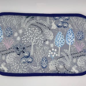 May include: A rectangular, padded item with a grey background featuring a whimsical forest scene. The design includes trees, rabbits, and birds in shades of white, blue, and pink. The item is trimmed with a dark blue border.