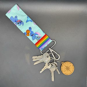Peut inclure: Un porte-clés bleu clair avec un motif de personnage de dessin animé, une bande arc-en-ciel et un fermoir argenté. Plusieurs clés et un charm en bois avec le texte "We all get a little lost. It's okay to there." sont attachés.