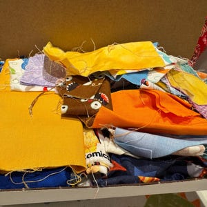 May include: A box filled with a variety of colourful fabric scraps. The scraps are in different sizes and patterns, including yellow, orange, blue, brown, and purple.