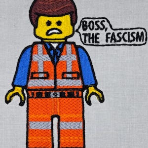 May include: An embroidered patch featuring a yellow plastic-style figure with brown hair, wearing a blue shirt and an orange high-visibility vest with silver reflective stripes. A speech bubble next to the figure contains the text BOSS, THE FASCISM.