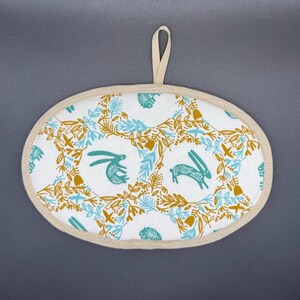 Peut inclure: Un dessous de plat ovale blanc avec une bordure beige et une boucle de suspension. Le tissu présente un motif répété de lapins turquoise et d'éléments floraux turquoise et dorés. Idéal pour la cuisine.