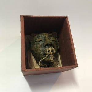 Könnte beinhalten: Ein geformter Kopf mit geschlossenen Augen und einem gequälten, gealterten Aussehen, präsentiert in einer braunen Box. Der Kopf hat eine grün-braune Farbgebung, wobei fadenartiges Material aus dem Mund ragt. Die Box ist mit einem beigen Stoff ausgekleidet.