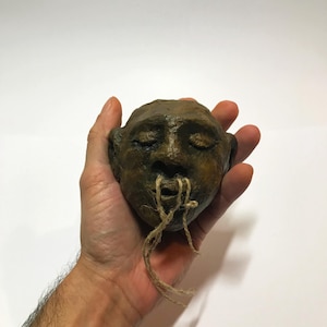 Könnte beinhalten: Kleine, strukturierte Skulptur eines menschlichen Gesichts mit geschlossenen Augen und neutralem Ausdruck. Das Gesicht hat eine gedämpfte braune Farbe, mit Schnüren, die aus dem Mund ragen. Die Skulptur wird in einer Hand vor weißem Hintergrund gehalten.