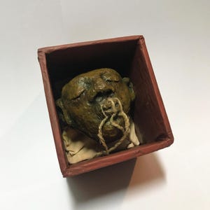 May include: A sculpted head with closed eyes, in shades of brown and green, rests inside a red box. A knotted rope, forming a heart shape, is attached to the head. The head is positioned on a beige cloth.