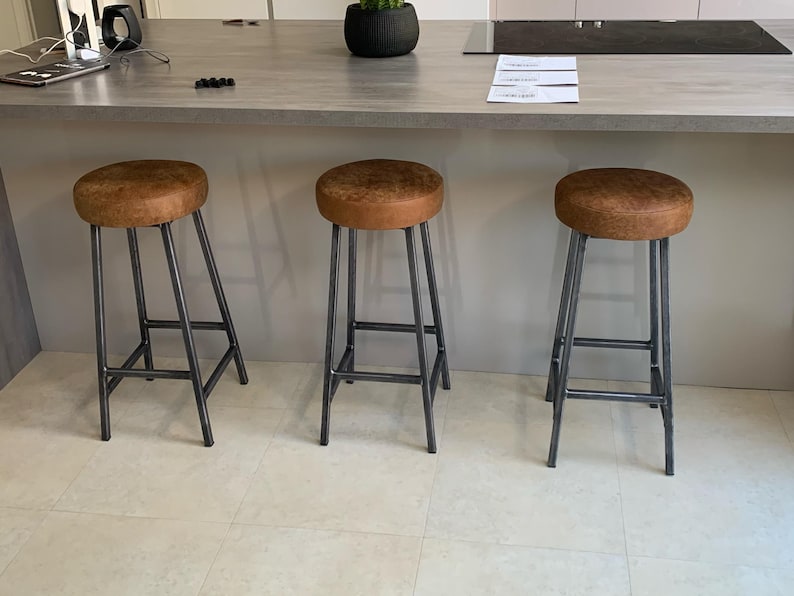 May include: Three brown leather bar stools with dark gray metal legs. The stools have a round seat and a simple, industrial design. The stools are positioned in front of a kitchen island.