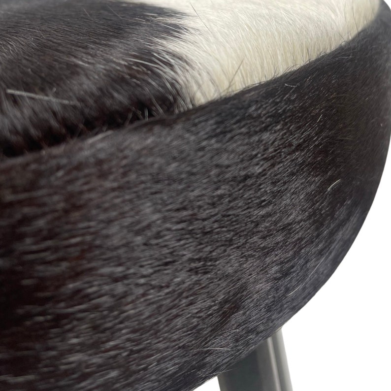 May include: Close-up of a black and white cowhide stool with a black base. The stool is made of soft, natural cowhide with a fuzzy texture.