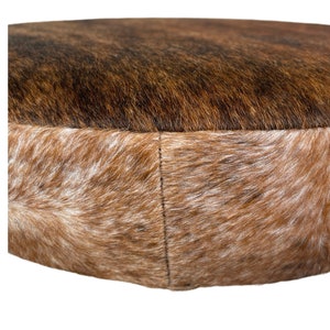 May include: A round brown and white cowhide ottoman with a stitched seam around the edge.