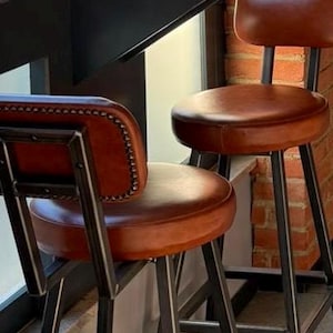 Op de afbeelding: Twee barkrukken met ronde, cognac-kleurige lederen zittingen en rugleuningen met metalen frames. De rugleuningen zijn aan de randen afgewerkt met een rij zilverkleurige siernagels. De krukken hebben zwarte, hoekige metalen poten en steunen.