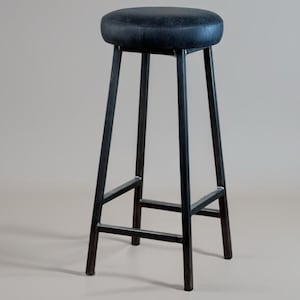 May include: A tall, black metal bar stool with a round, padded seat. The seat is a dark, textured material. The stool has four legs that angle outward for stability, connected by horizontal supports.