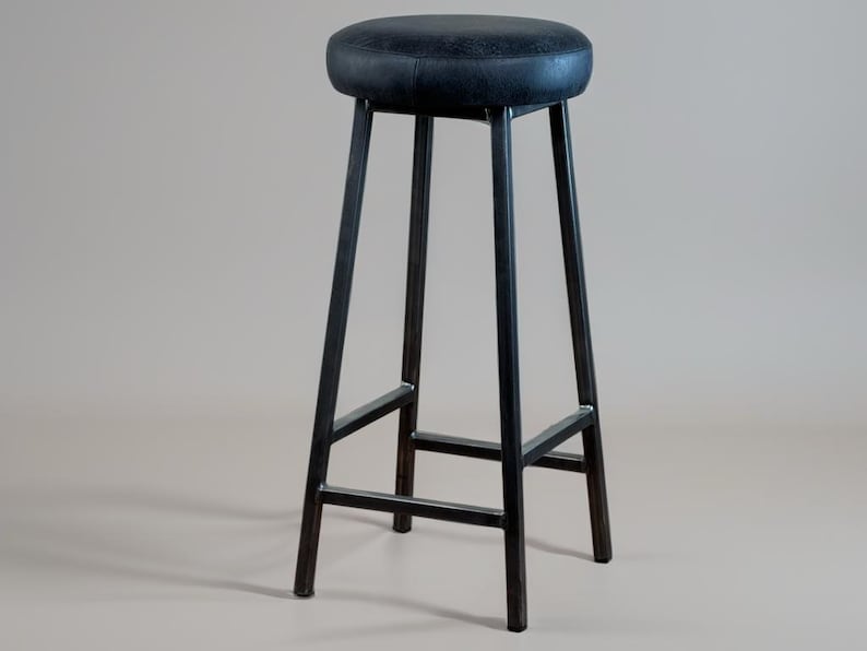 May include: A tall, black metal bar stool with a round, padded seat. The seat is a dark, textured material. The stool has four legs that angle outward for stability, connected by horizontal supports.