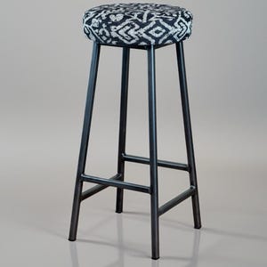 May include: A tall bar stool with a round seat and a black metal frame. The seat features a black and white geometric pattern. The stool's legs are angled and connected by horizontal supports, creating a sturdy base. The stool is designed for a bar or counter.