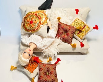Könnte beinhalten: Eine Puppe mit langen blonden Haaren liegt auf einem weißen Sofa mit mehreren dekorativen Kissen. Die Kissen sind mit roten, goldenen und orangefarbenen Mustern bestickt.