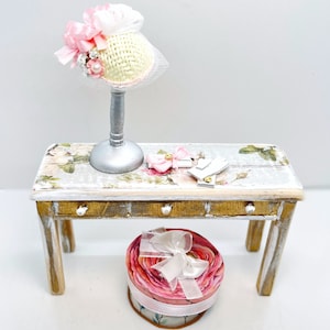 May include: A white and pink hat with a white veil sits on a silver stand on a white and gold table with a pink and white floral design. A pink and white gift box sits on the table.