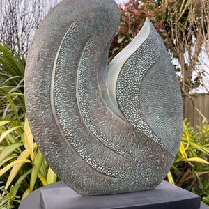 Könnte beinhalten: Eine grüne Bronzeskulptur mit abstrakten Formen und einer strukturierten Oberfläche. Die Skulptur steht auf einem schwarzen Sockel.