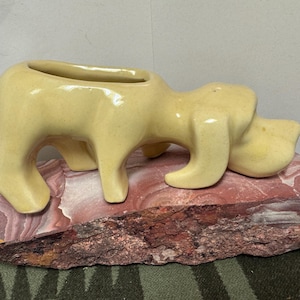 May include: A ceramic figurine of a yellow hippo-like animal, resting on a reddish-brown stone base. The figurine has a hollowed-out back, suggesting it could be a planter or decorative item. The stone base has a rough, natural texture.