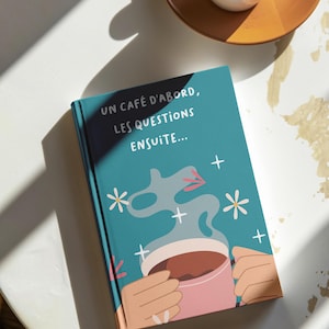 Peut inclure: Un carnet bleu turquoise avec une illustration blanche et rose d'une main tenant une tasse de café fumante. La couverture du carnet porte le texte "Un café d'abord, les questions ensuite..." en blanc.