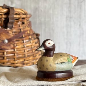 May include: A small, hand-painted wooden duck figurine featuring a dark brown head with a white eye patch, a tan body with brown speckles, and light blue and orange wing details, mounted on a circular dark brown wooden base with a felt bottom.