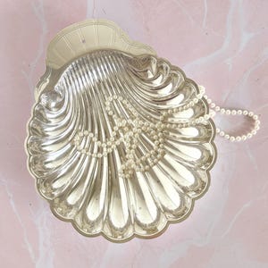 May include: A silver-toned metal dish shaped like a large scallop shell with a scalloped edge and a small hole at the top for hanging. A single strand of white pearl beads is draped across the interior of the shell, resting on the curved ridges.