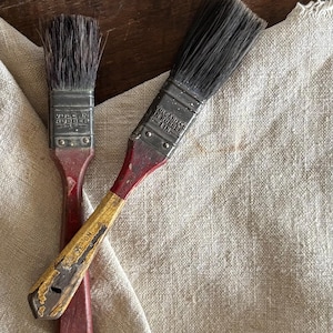 May include: Two vintage paint brushes with dark bristles and metal ferrules stamped with VULCANIZED IN RUBBER. The smaller brush has a solid red handle, while the larger brush features a worn yellow and red handle with the text NO. 35 visible.
