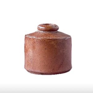 May include: A small, cylindrical, brown ceramic jar with a lid. The jar has a rounded top and a small, circular knob on the lid. The exterior has a textured surface and a rustic appearance. The jar is likely for storage.