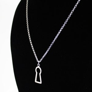 May include: A silver-colored necklace featuring a keyhole pendant. The pendant is a simple, open design, suspended from a delicate chain. The necklace is displayed against a black background.