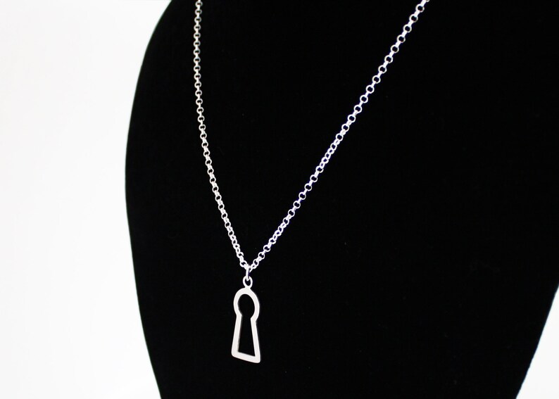 May include: A silver-colored necklace featuring a keyhole pendant. The pendant is a simple, open design, suspended from a delicate chain. The necklace is displayed against a black background.